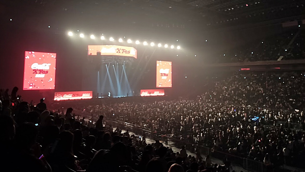 K-POPコンサート会場の広大なアリーナ席。多くの観客がステージに向かって立ち上がり、ペンライトを振って熱心に応援する光景。