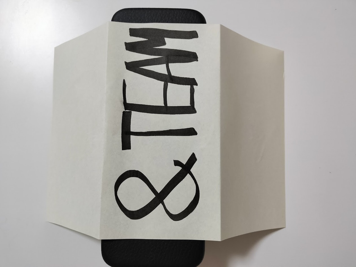 眼鏡ケースに「&TEAM」と書いた紙を巻き付けて、お菓子の巻き紙のサイズ感を確認している様子